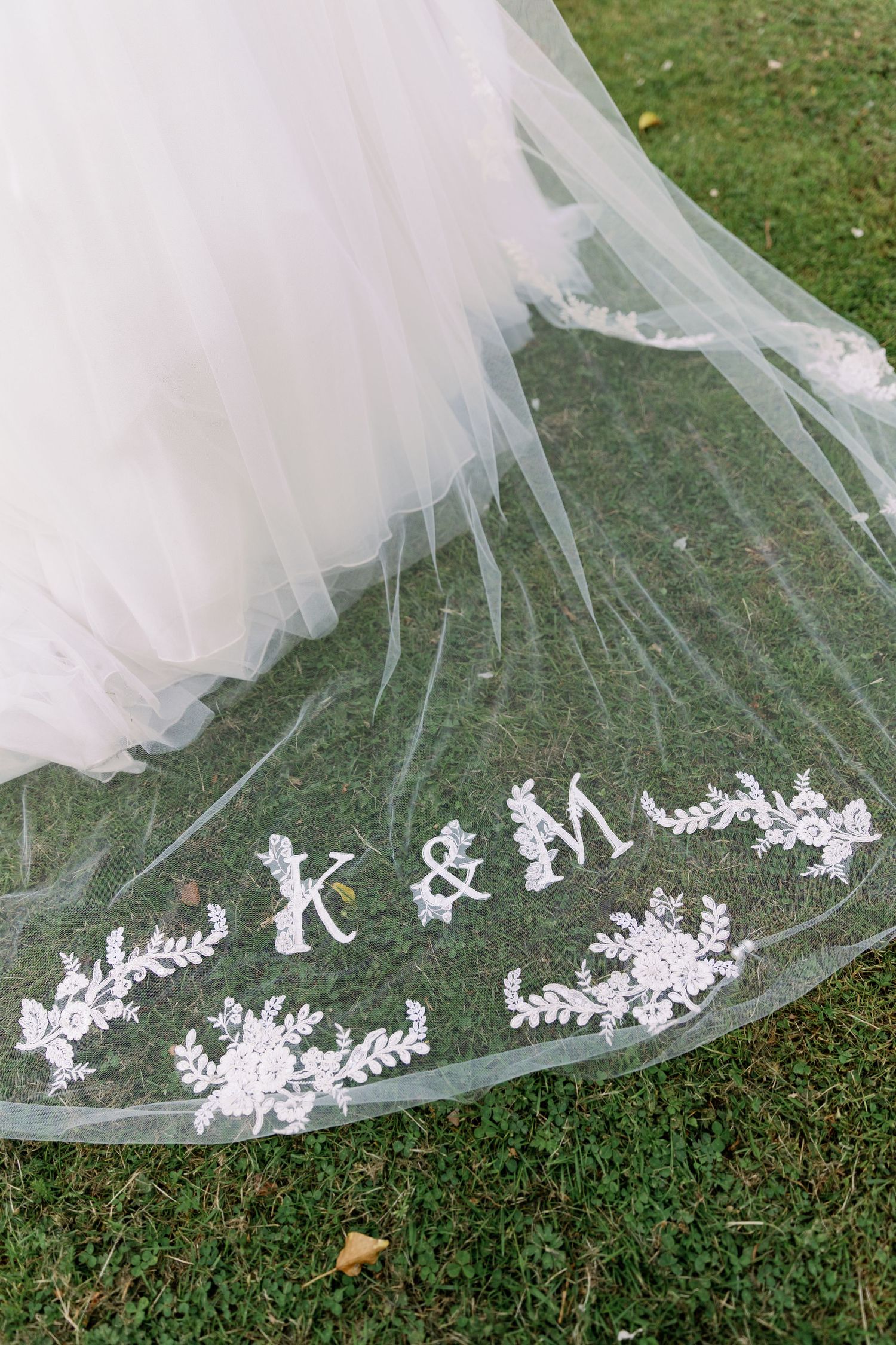 Wedding veil with delicate white lace embroidery and monogram initials K & M laid out on green grass.