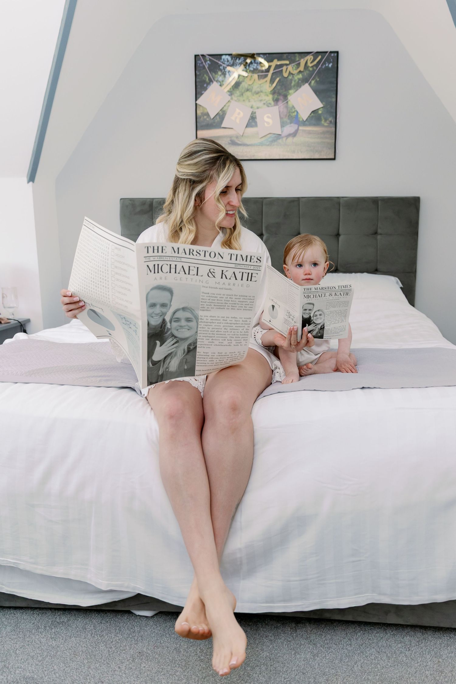 Woman and baby sitting on white bed reading a newspaper together while relaxing at home.