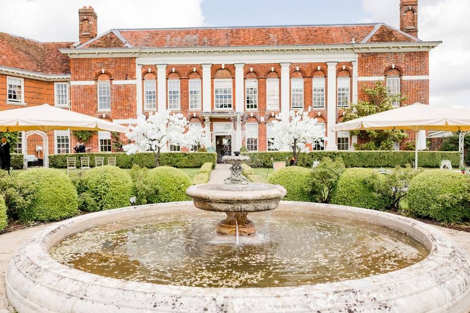 Elegant brick mansion with grand colonnade facade and ornate water fountain in manicured formal garden courtyard.