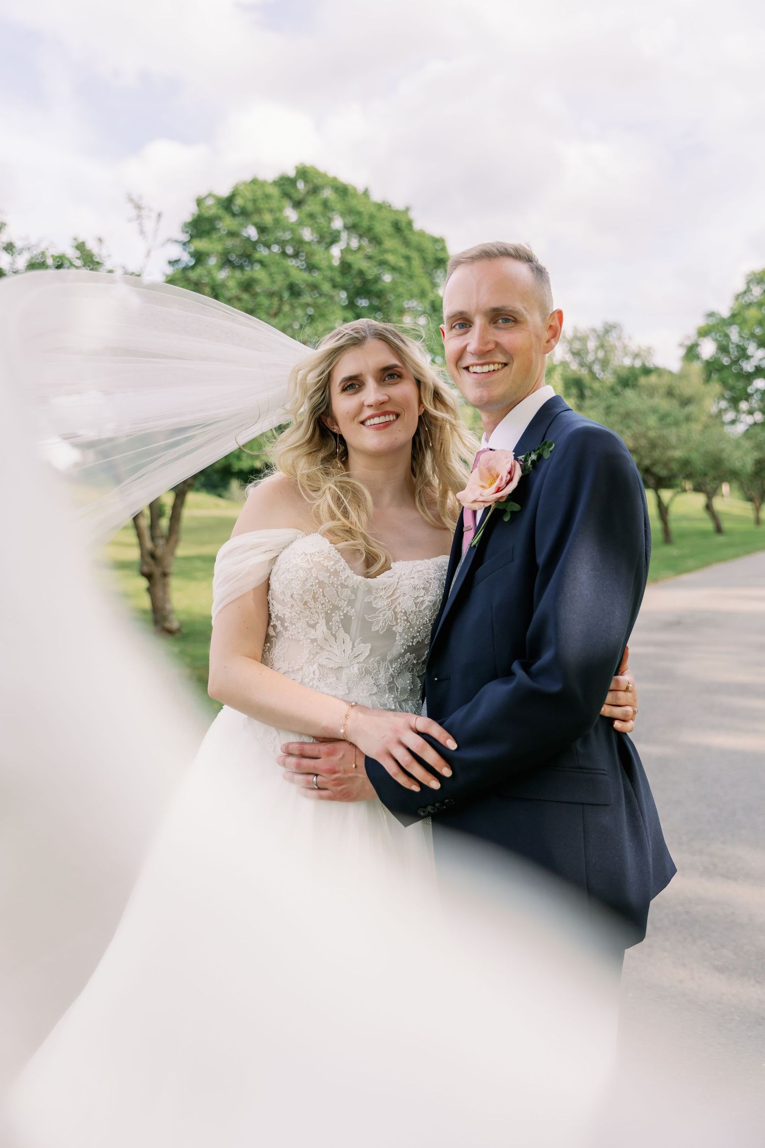 A happy couple on their wedding day stand together outdoors with a flowing veil catching the breeze.