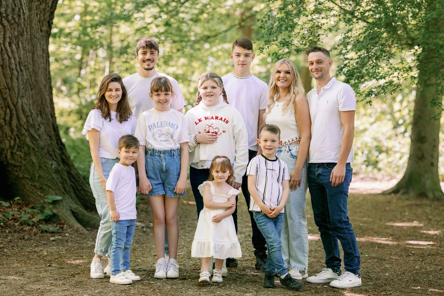 A group of people in casual white and denim outfits poses together for a photo near a large tree in a forest setting.