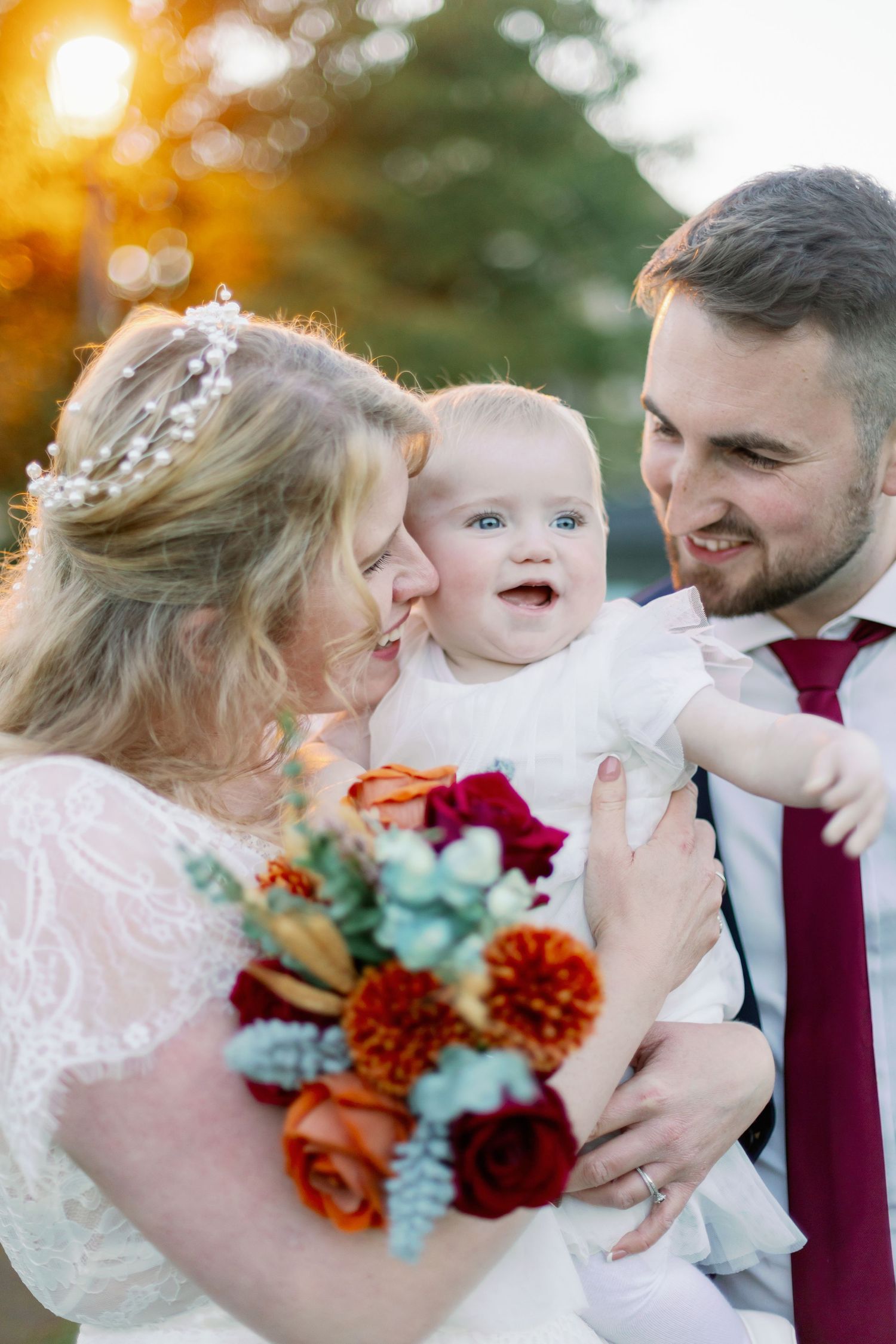 Beautiful autumn wedding photo outdoors with happy family holding vibrant red and orange bouquet in sunlit garden setting.