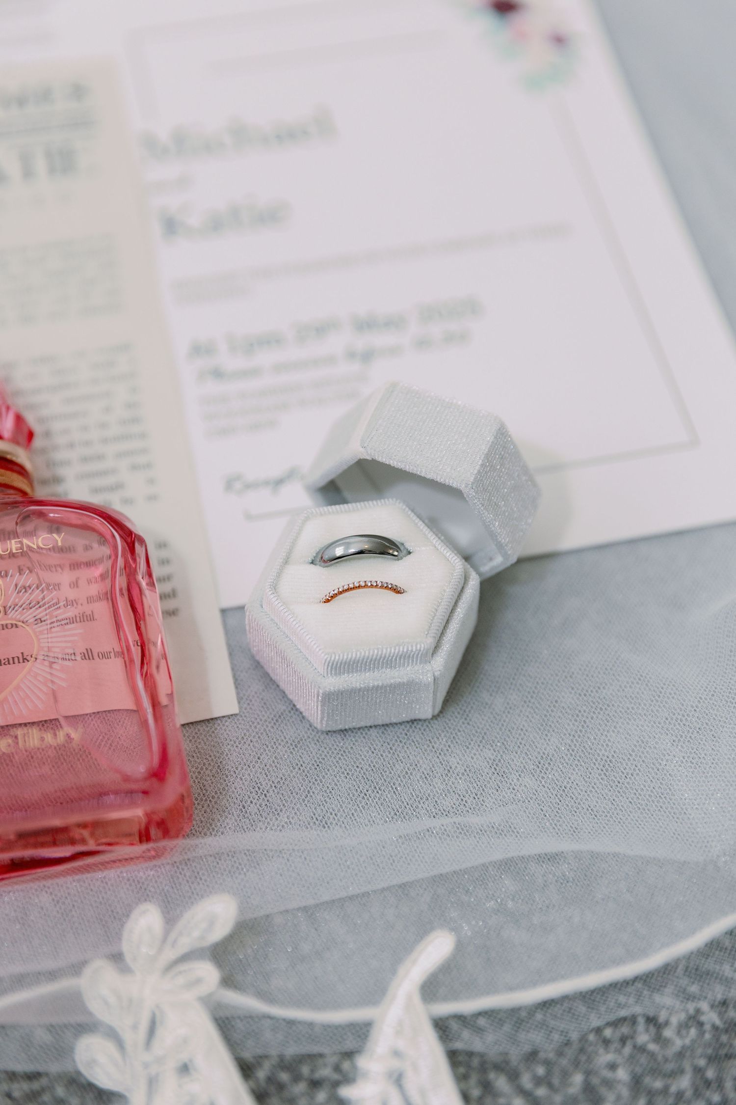 A wedding ring in a white hexagonal box next to a pink perfume bottle and paper documents.