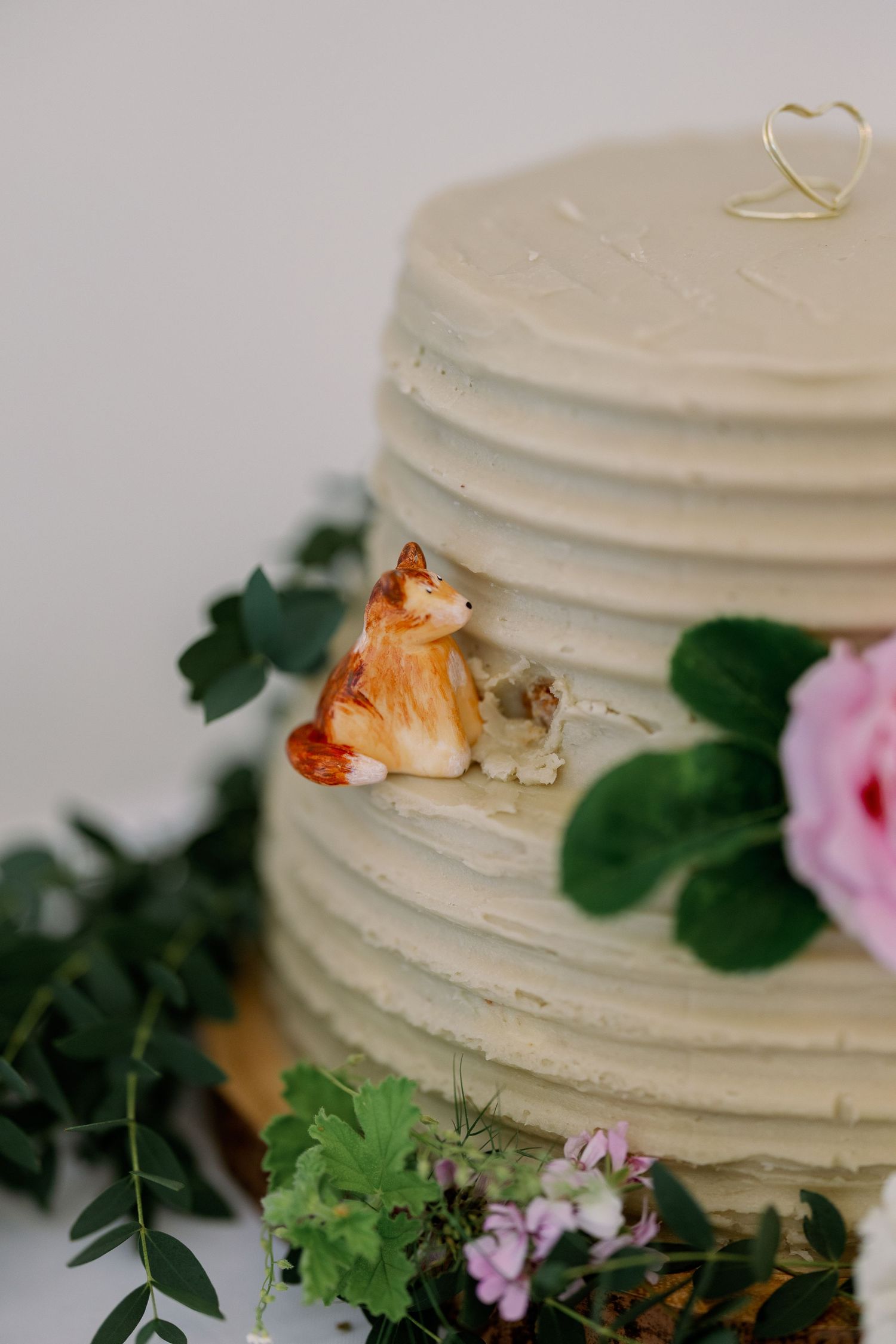 White tiered wedding cake decorated with a small ceramic cat figurine and pink flowers with green foliage.