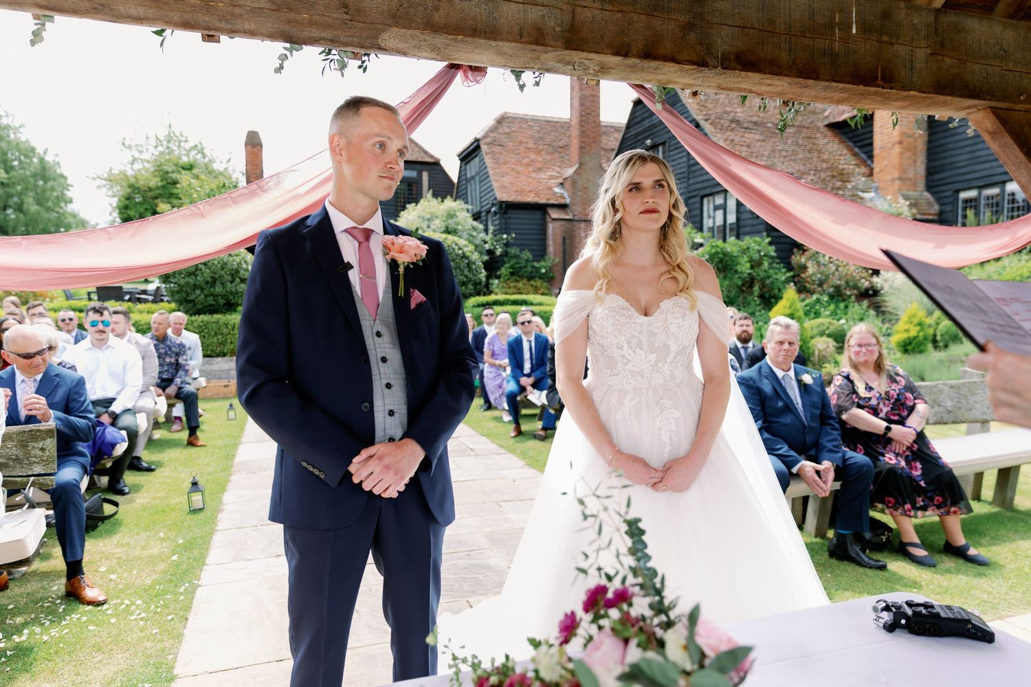 An outdoor wedding ceremony under a rustic wooden pavilion with a couple standing before seated guests in a garden setting.