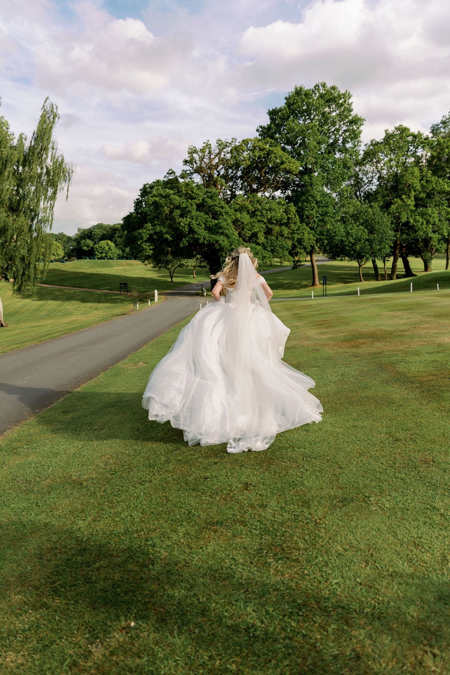 A flowing white wedding dress billows in the breeze as someone walks along a paved path on a lush golf course.