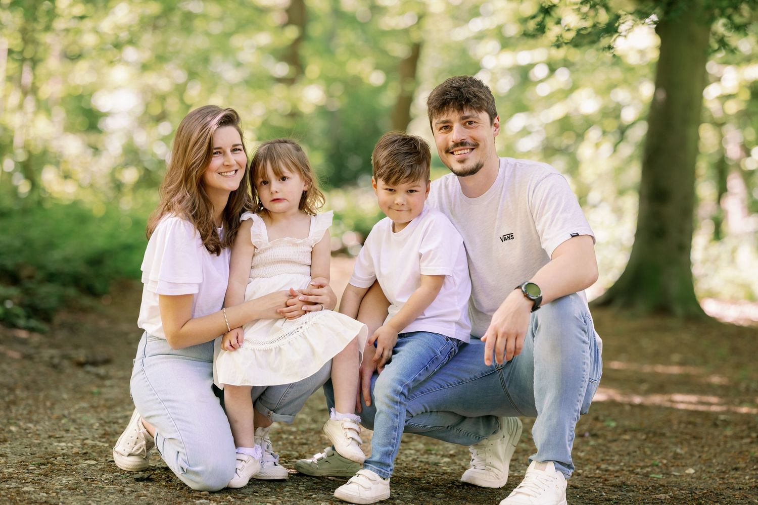 Three people in white clothing kneel together in a sunlit forest setting while enjoying a casual outdoor moment.