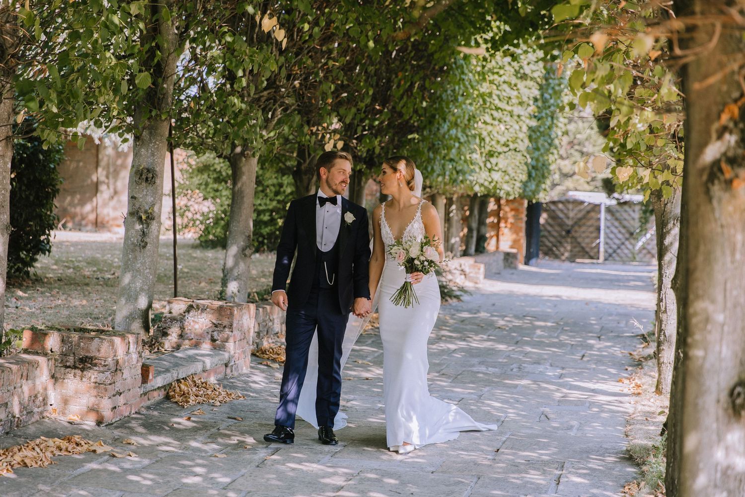 A newlywed couple walks together under tree-lined walkway holding hands on their wedding day.
