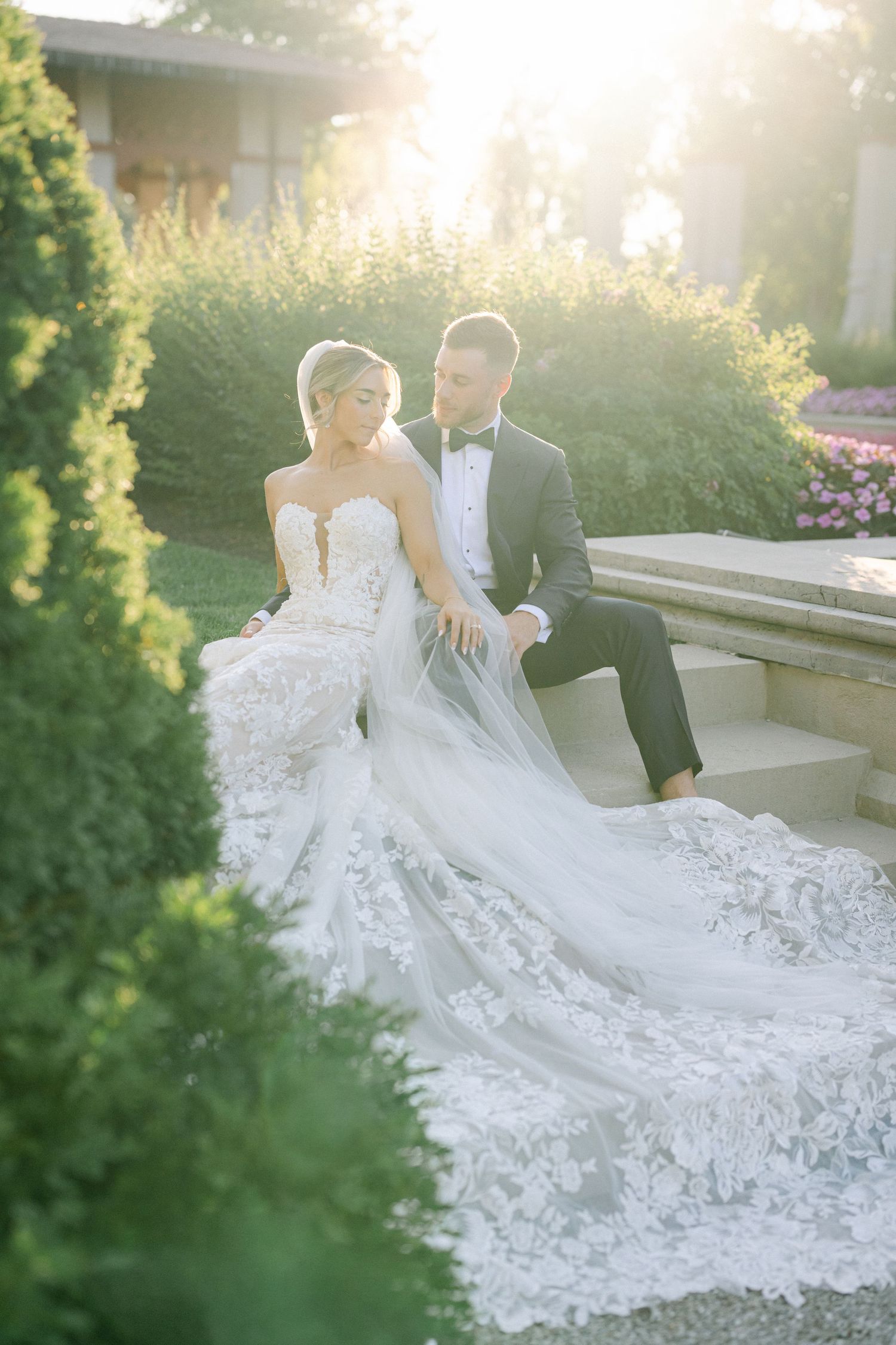 A romantic wedding couple sits on stone steps during golden hour as the bride's lace dress flows elegantly beside them. - The Armour House