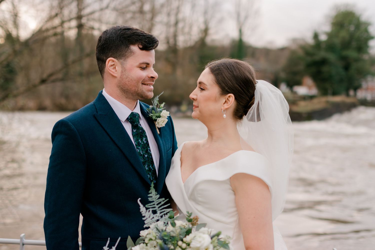 A happy couple poses together against a wintry outdoor backdrop holding a wedding bouquet with blue and white flowers.
