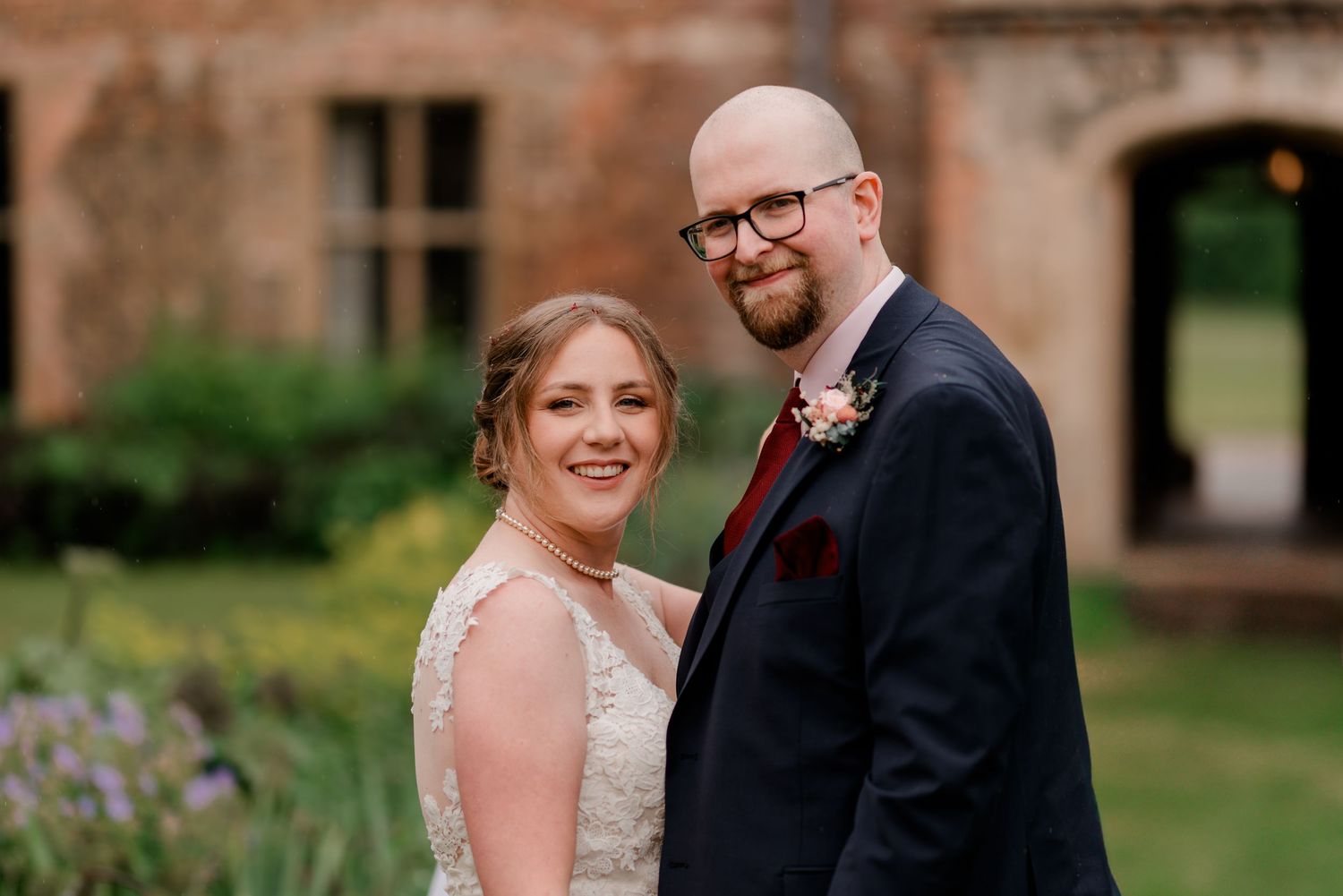 Romantic wedding portrait showing couple in elegant attire embracing outdoors against rustic brick and garden scenery.