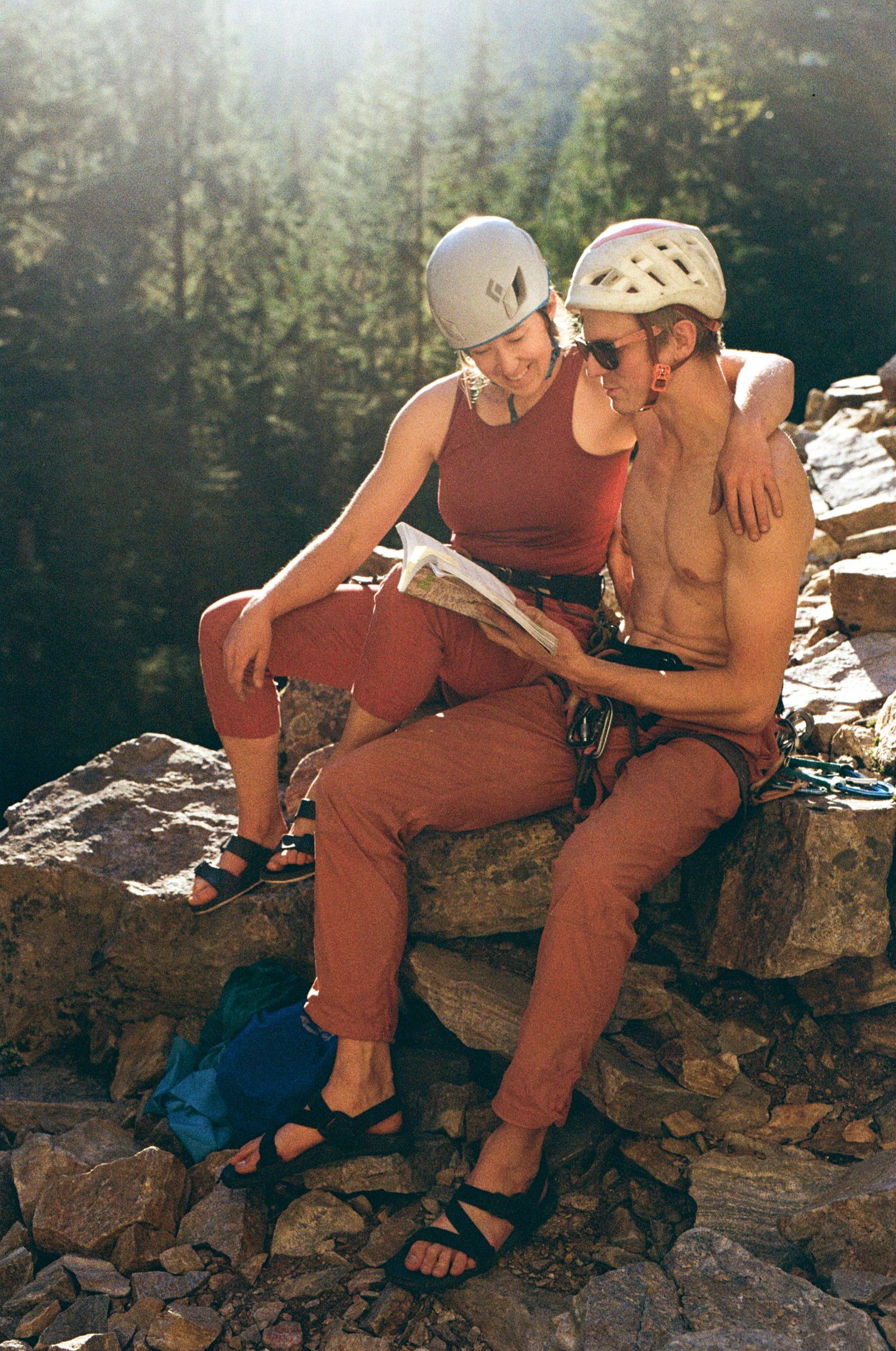 couple in rock climbing gear cuddle and look at a guide book with the sun shining behind them in Revelstoke, BC