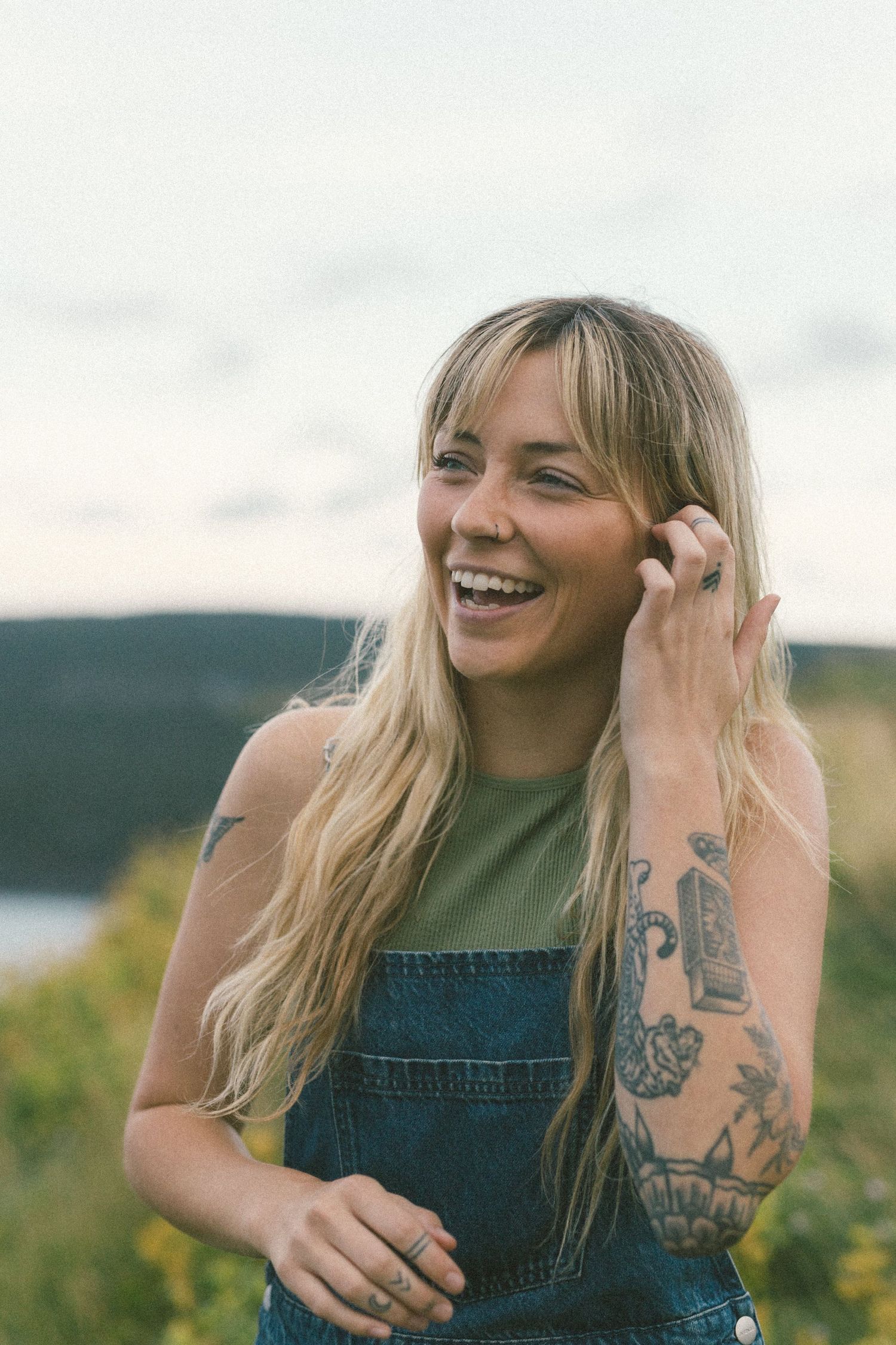blonde woman with tattoos laughs and tucks her hair behind her ear in a field of tall grass during Self Love Session