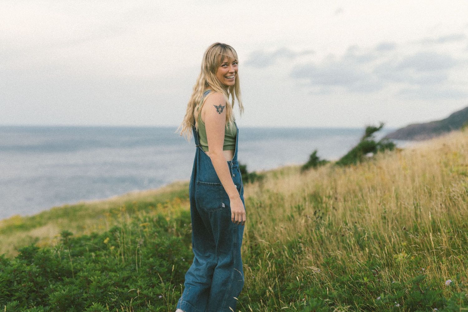 blonde woman with tattoos laughs in a grassy meadow above the ocean