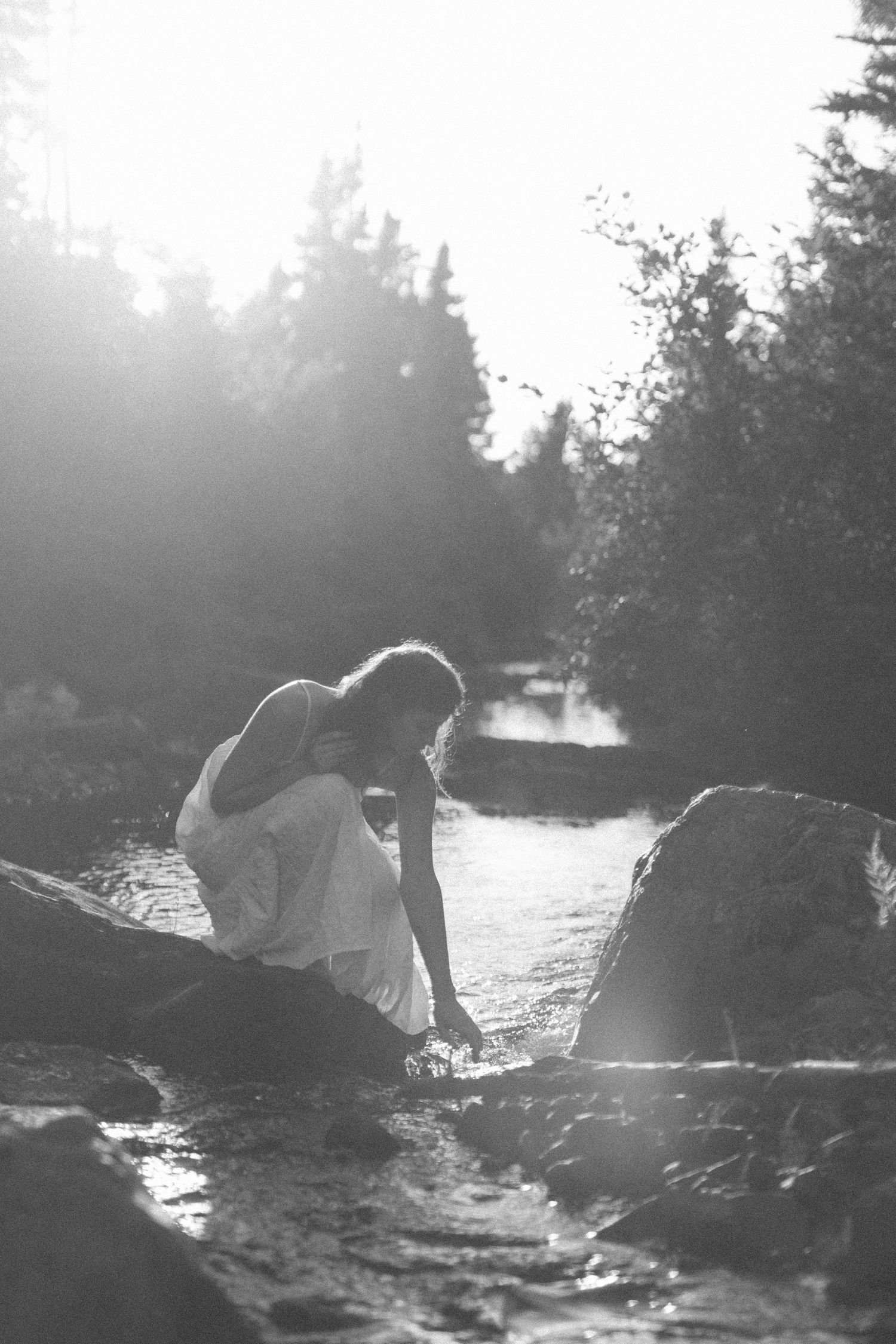 woman in a long white dress bends down to dip her fingers in a river during self love session