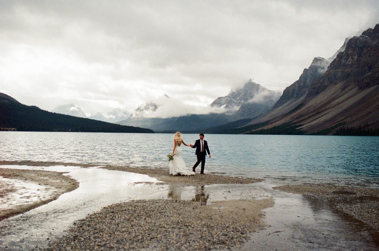 35 mm film photo of bride and groom walking along shore of Bow Lake in Banff with mountains towering above them