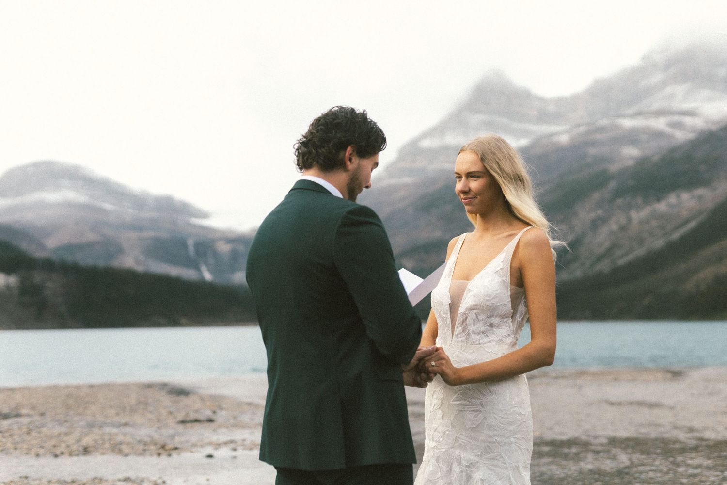 bride smiles at groom as he reads his vows at Bow Lake in Banff National Park