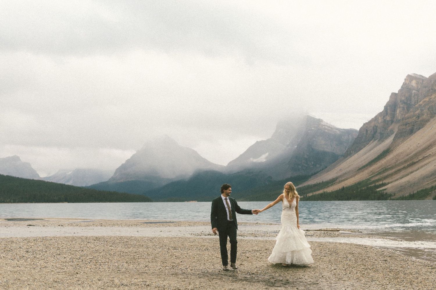 35 mm film photo of bride and groom walking along shore of Bow Lake in Banff with mountains towering above them