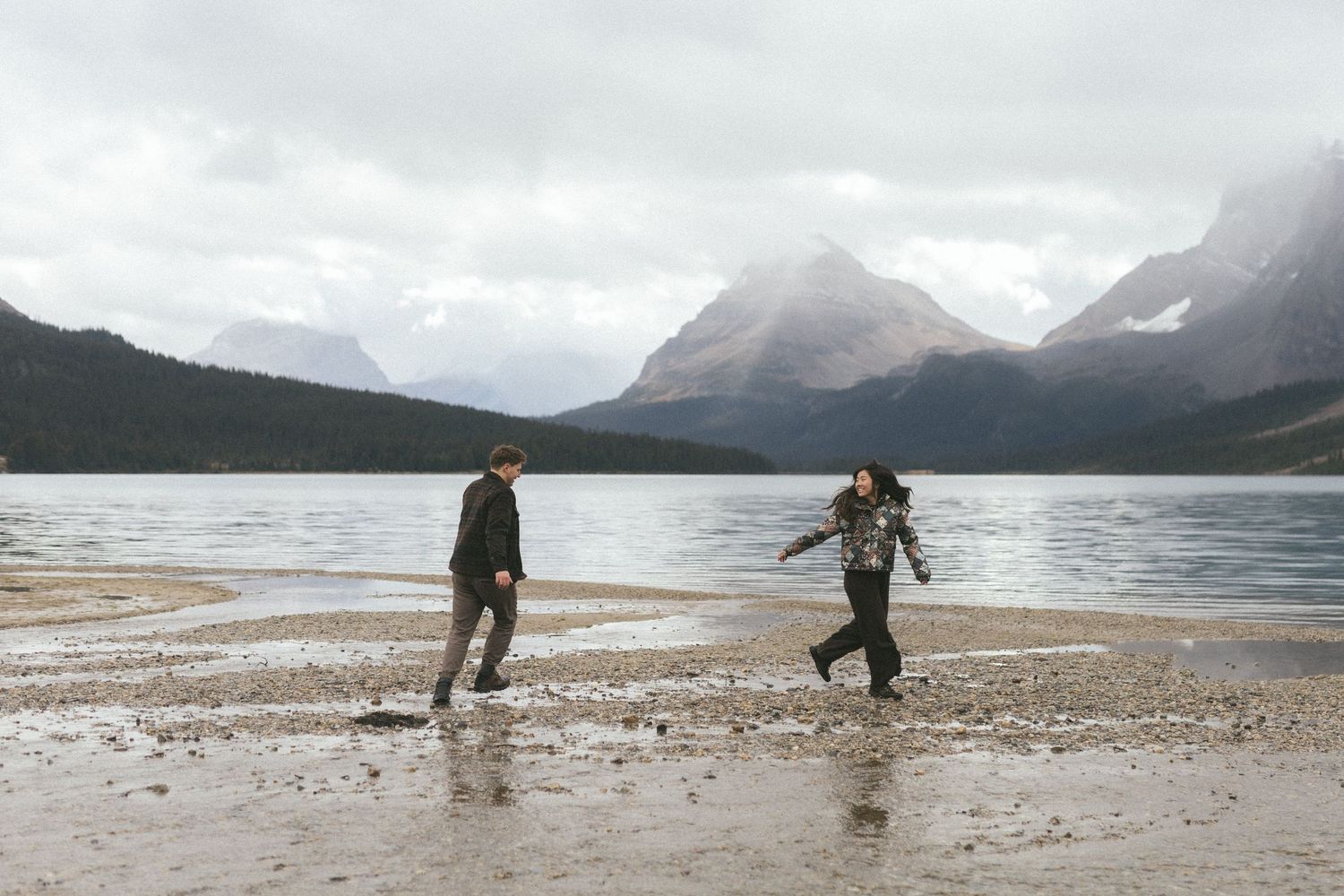 couple run along beach at Bow Lake in Banff with mountains behind them