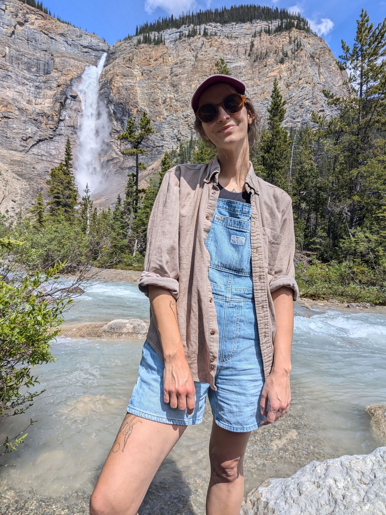 Tofino Elopement Photographer Heather Nolan smiles serenely at the camera in overalls and flannel standing beside Takkakaw Falls in Yoho
