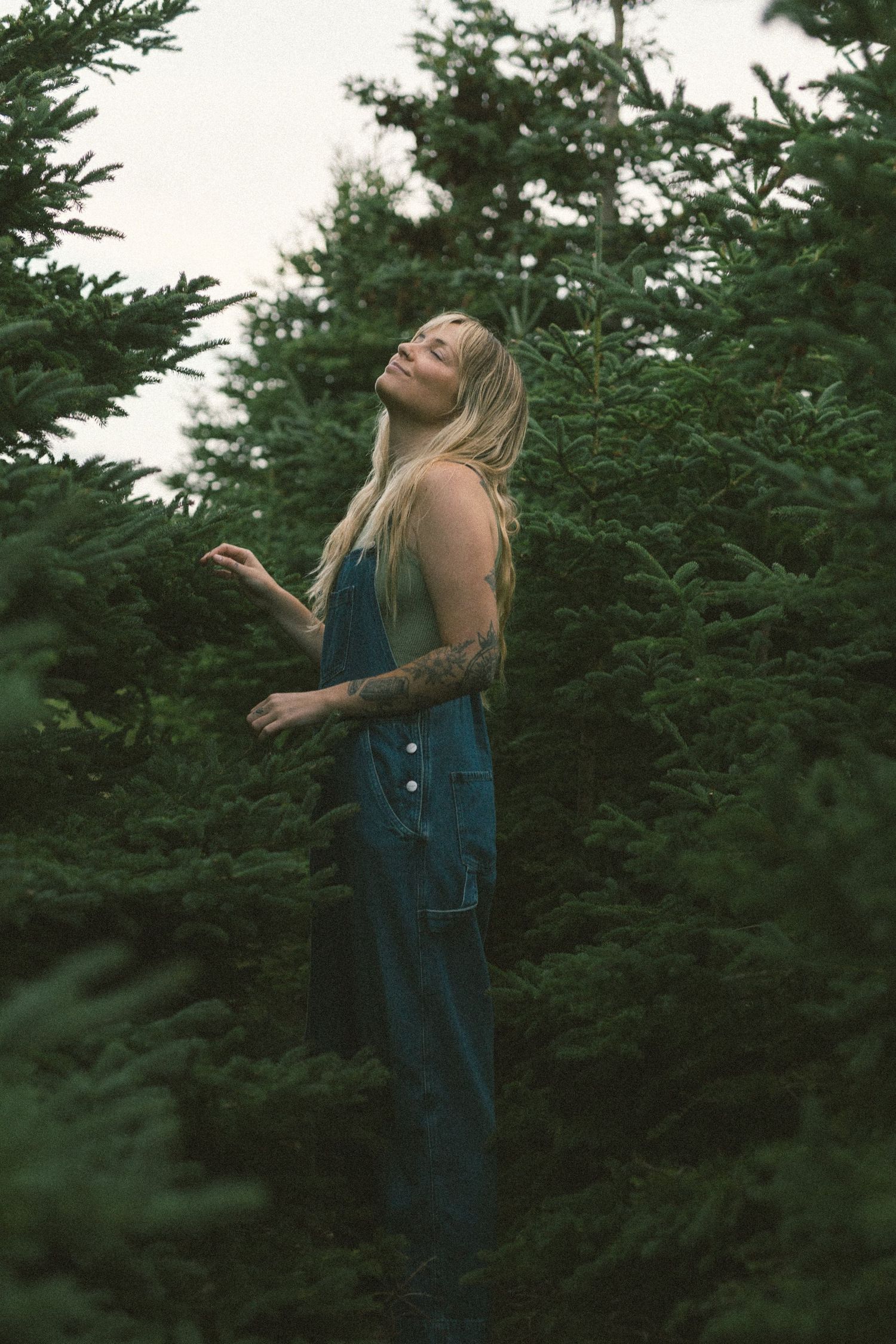 white woman with tattoos closes her eyes and takes a deep breath of the spruce trees she is surrounded by