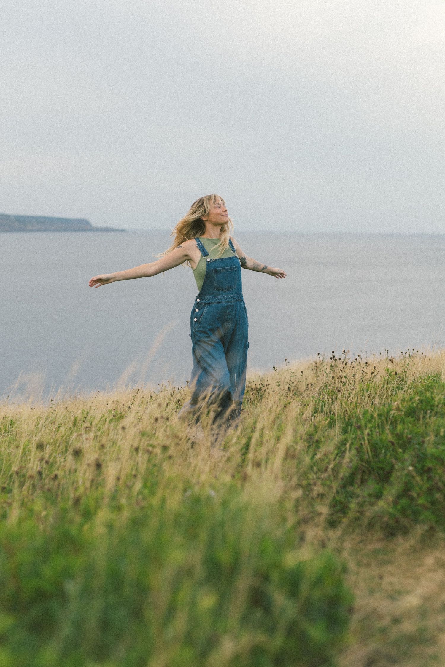 blonde woman with tattoos spins around in a meadow by the ocean during Self Love Session