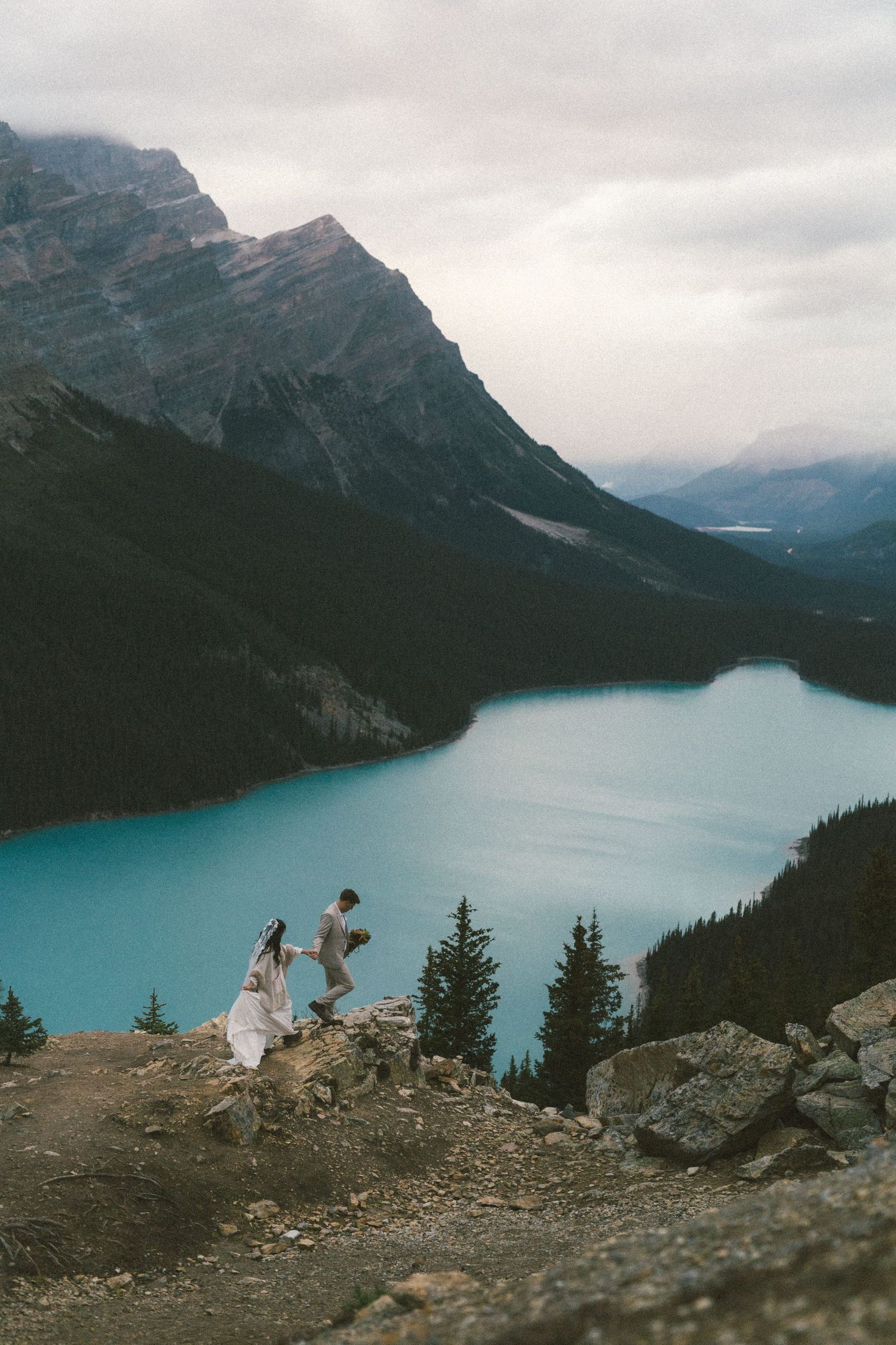 bride and groom walk along ridge line above a mountain lake in Banff National Park