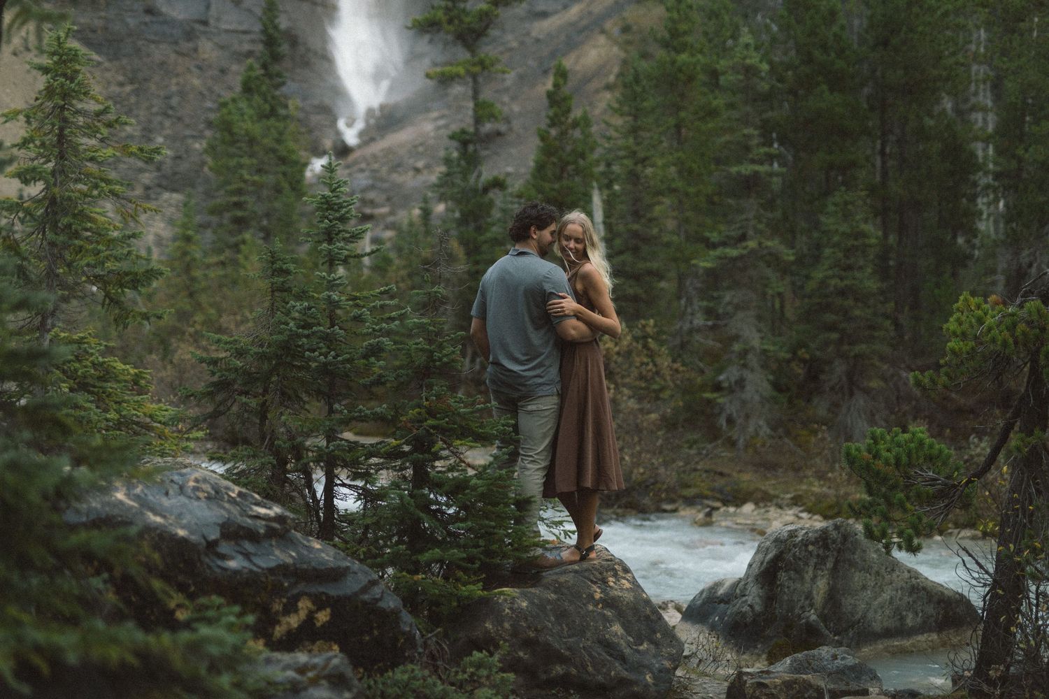 couple hold each other and laugh beside Takkakaw Falls in Yoho National Park