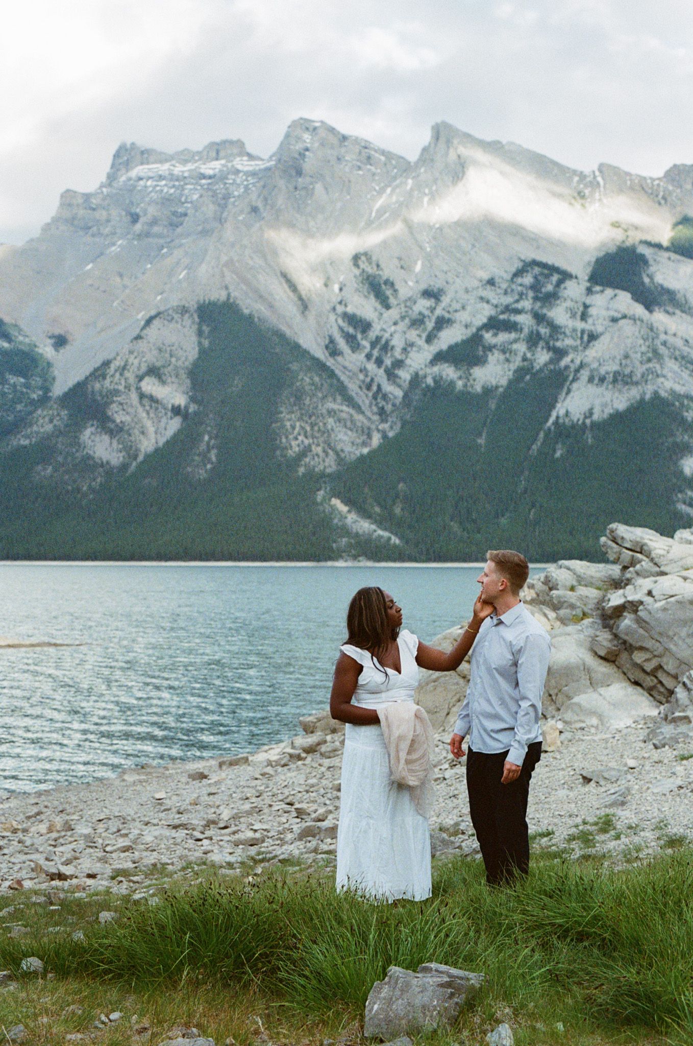 35 mm film photo of bride touching groom's face on the shore of Lake Minnewanka in Banff with mountains rising above them