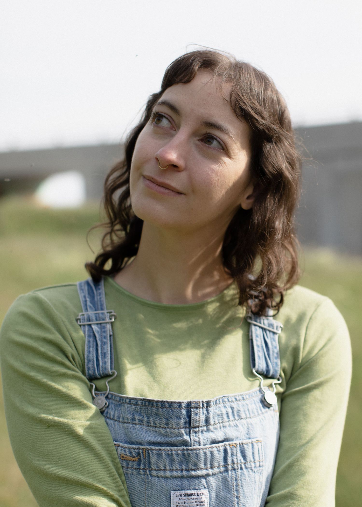 Banff Elopement Photographer Heather Nolan smiles serenely as they look off to the left. They are wearing denim overalls and a green shirt