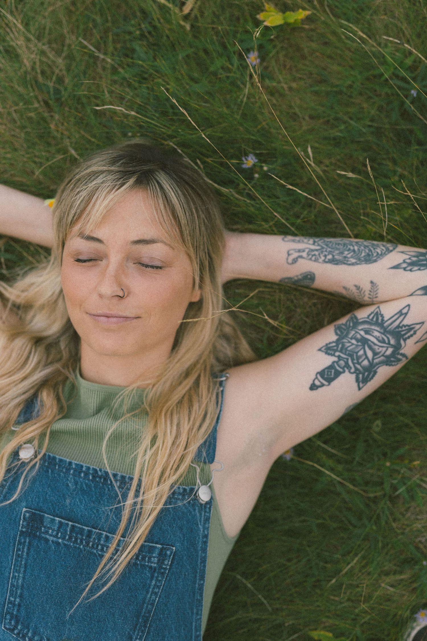blonde woman with tattoos lays in the tall grass with her eyes closed and a peaceful smile during Self Love Session