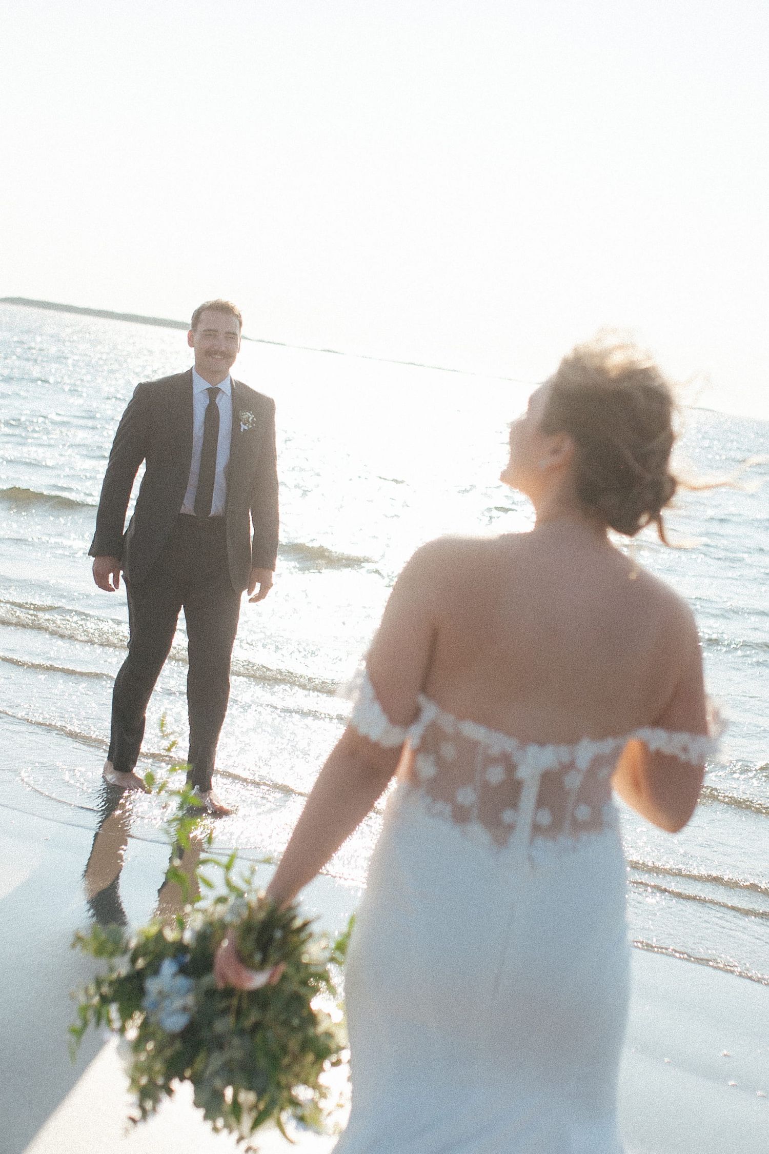 bride and groom face each other laughing in the glare of the sun with gentle waves around them in Tofino