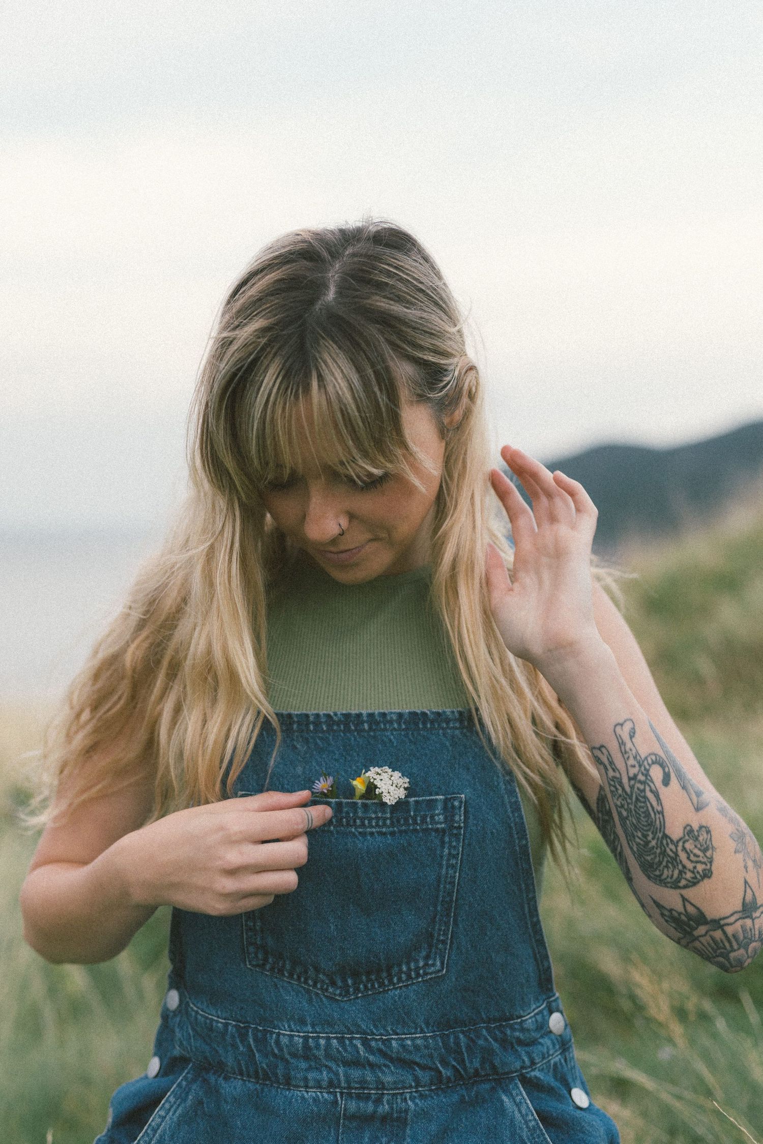 blonde woman with tattoos puts wildflowers in her overalls pocket