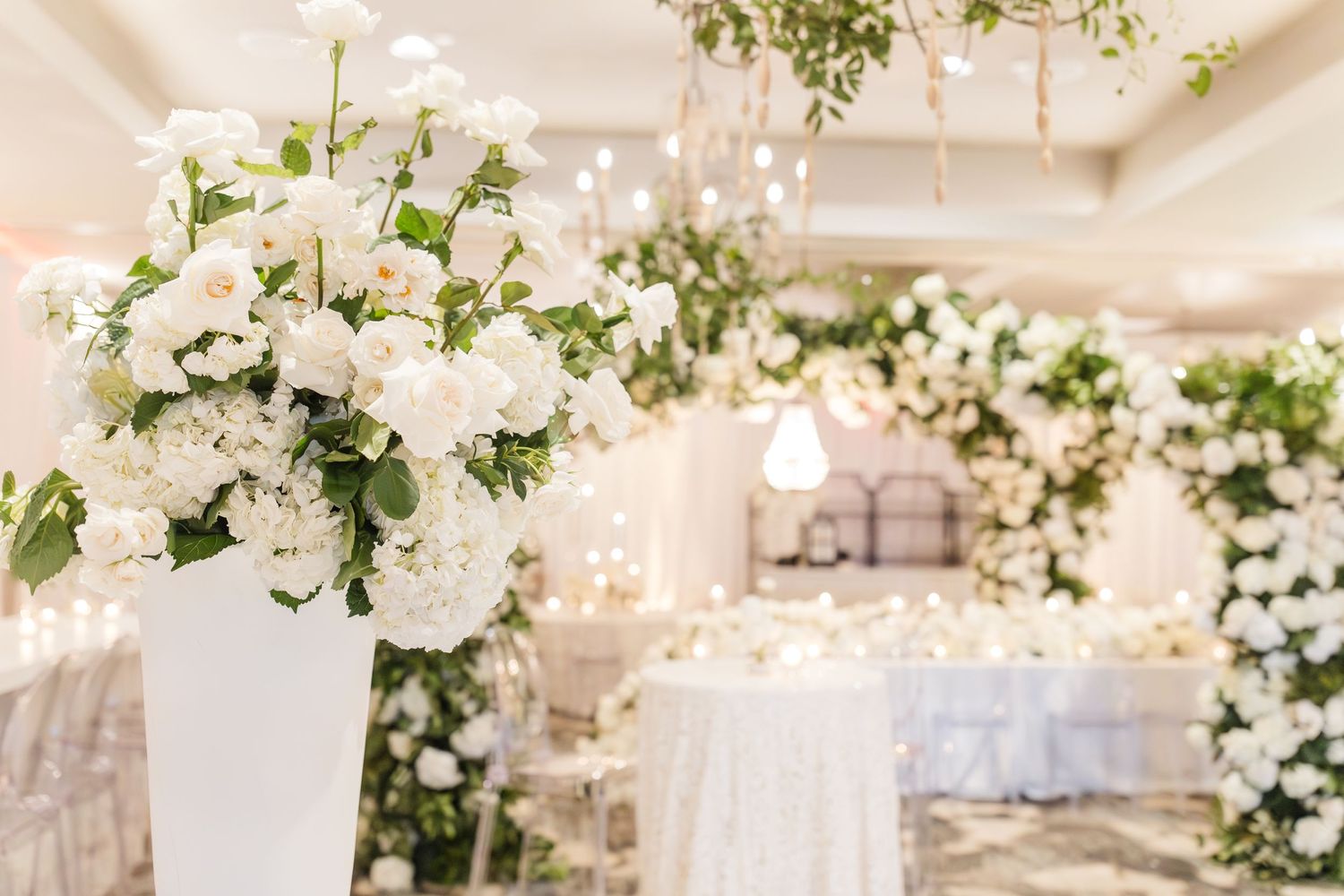 Elegant white floral arrangements and arch decorations adorn a luxurious indoor wedding reception venue with soft lighting.