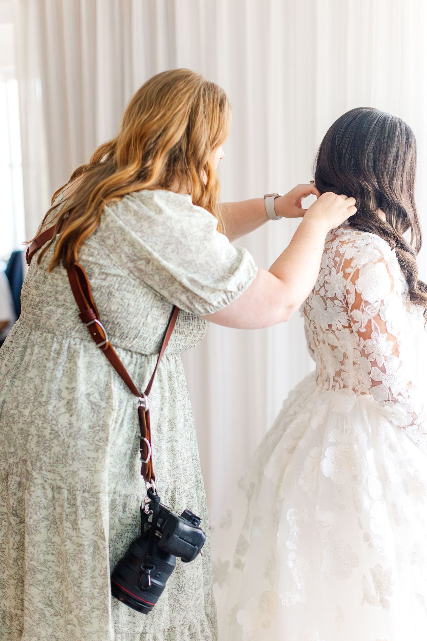 Wedding photographer with camera adjusts bride's hair while she wears a white floral gown against light curtains.