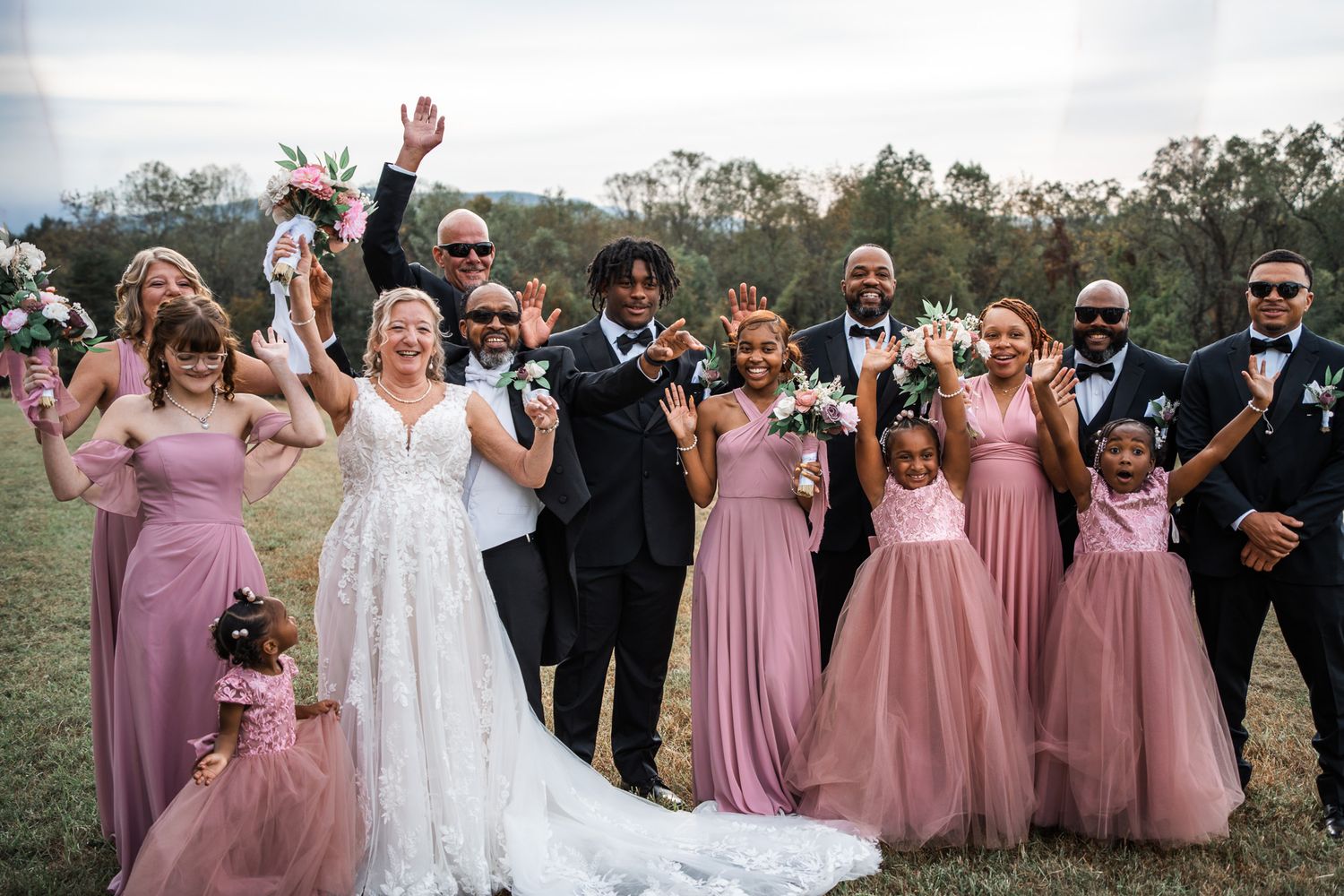 Wedding party in pink bridesmaid dresses and black suits pose together outdoors on a cloudy day.
