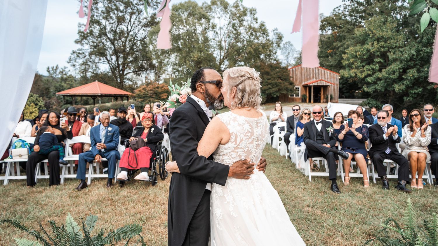 A couple shares their first dance at an outdoor wedding ceremony with guests seated on white chairs in the background.