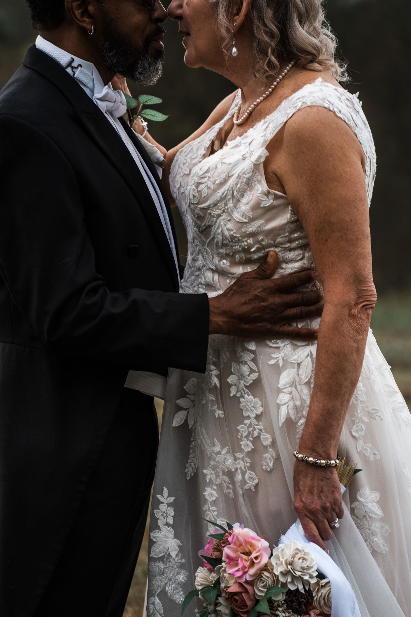 Close-up shot of a bride in a lace wedding dress and groom in black suit sharing an intimate moment with pink bouquet.