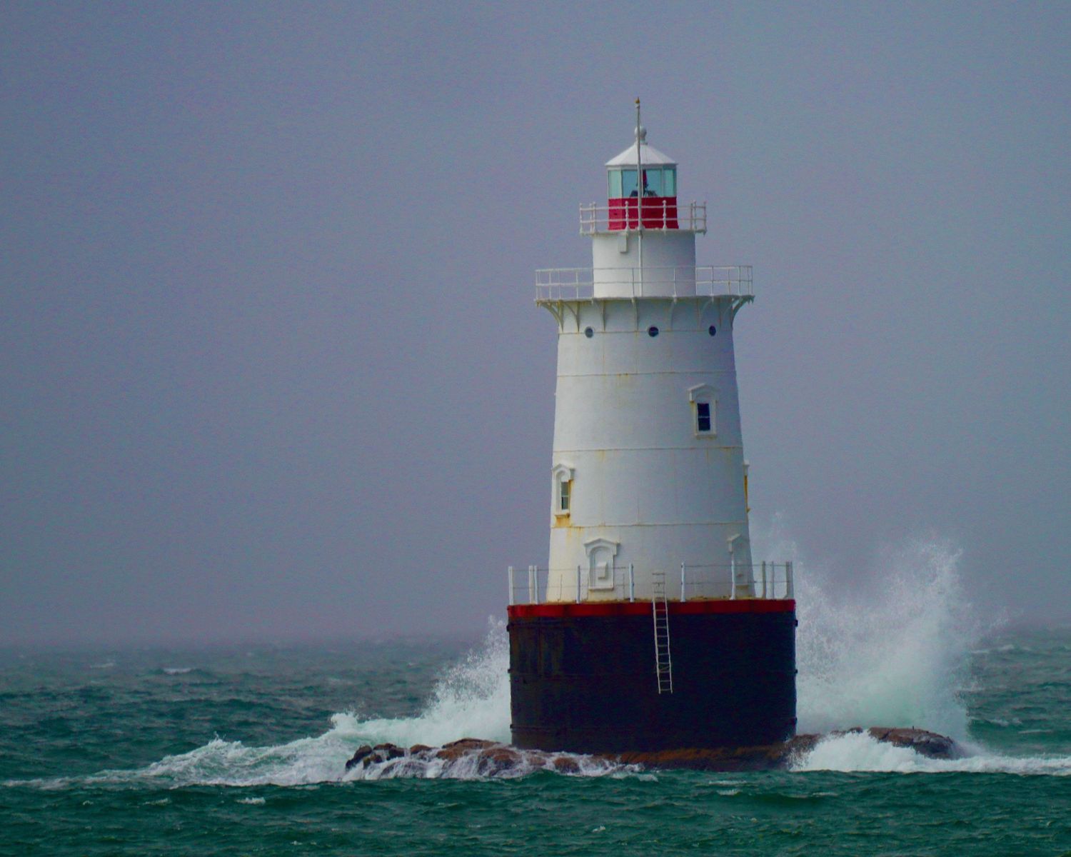 Waves crash against a white and red lighthouse in stormy seas with dark green waters and gray skies.