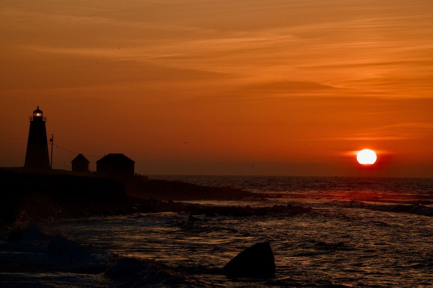 Dramatic orange sunset silhouettes a lighthouse and rocky coastline against a vibrant sky.