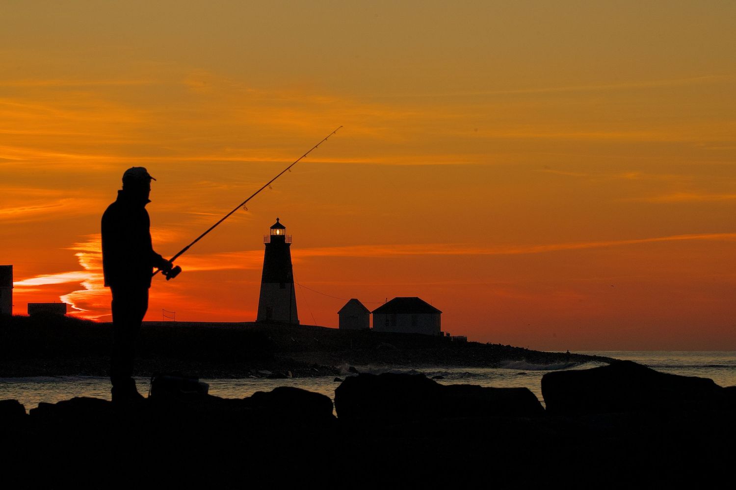 Silhouettes of fishing figures and lighthouse against a brilliant orange sunset over the ocean.