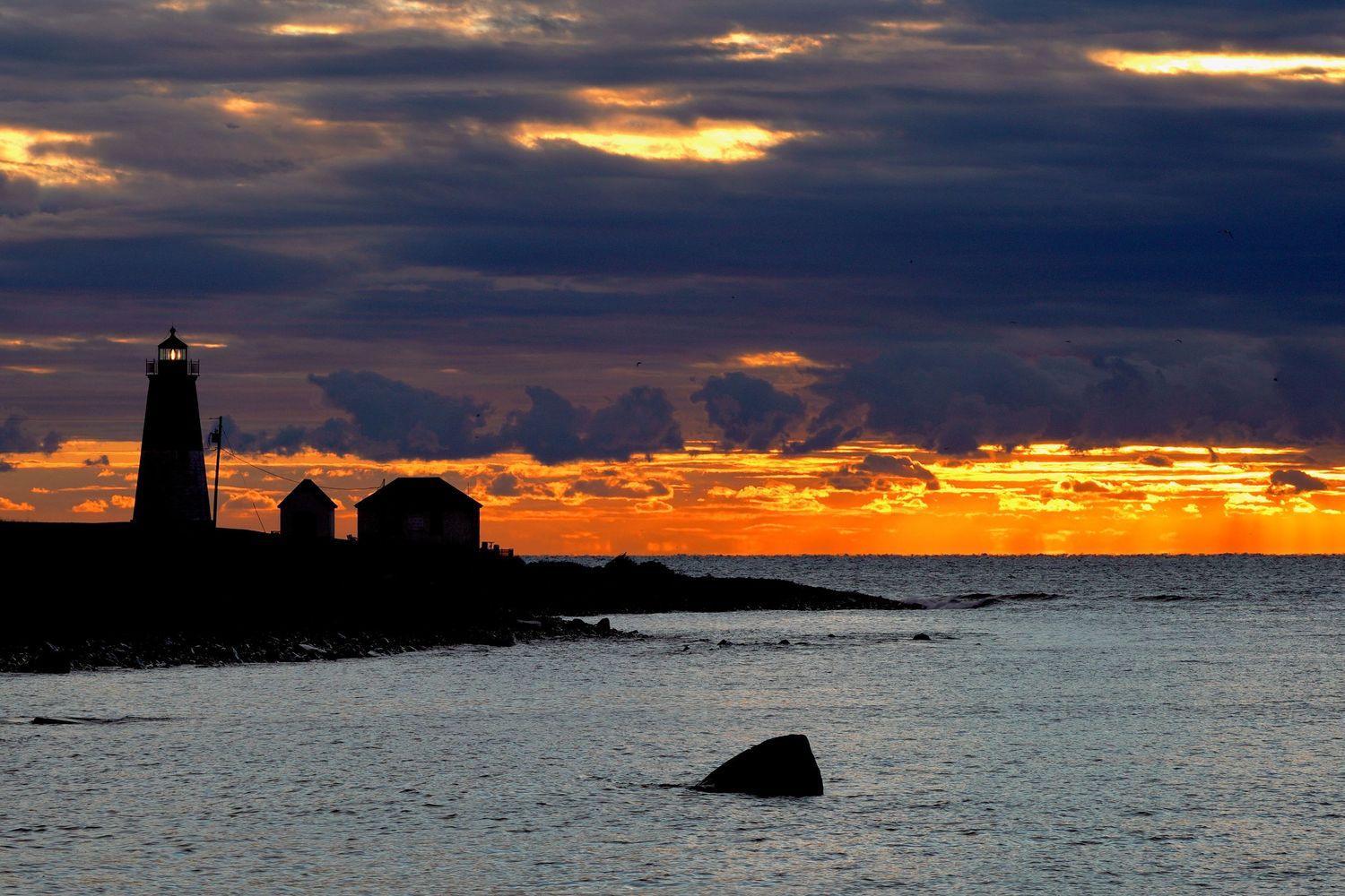 Lighthouse stands sentinel against fiery sunset clouds over the ocean horizon.