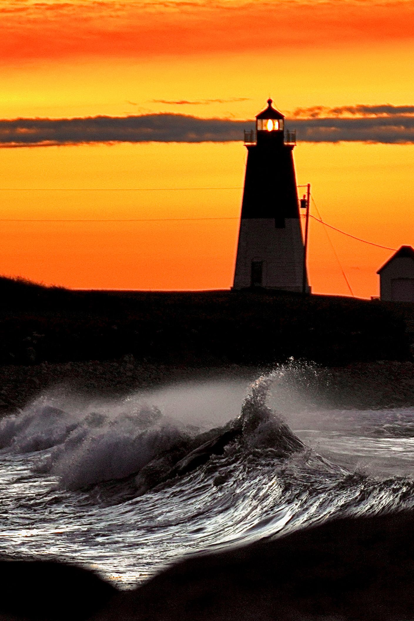 Dramatic ocean waves crash against a lighthouse silhouette during a vibrant orange and yellow sunset.