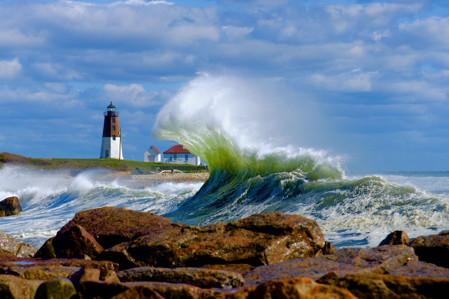 A massive green wave crashes against rocky shores near Point Judith Lighthouse in Rhode Island on a sunny day.