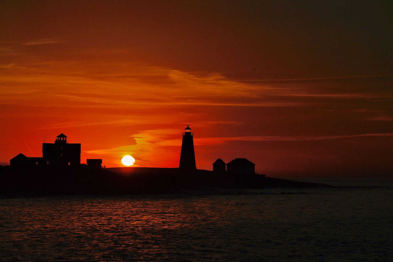 Dramatic silhouette of buildings against a vibrant orange sunset sky over the ocean horizon.