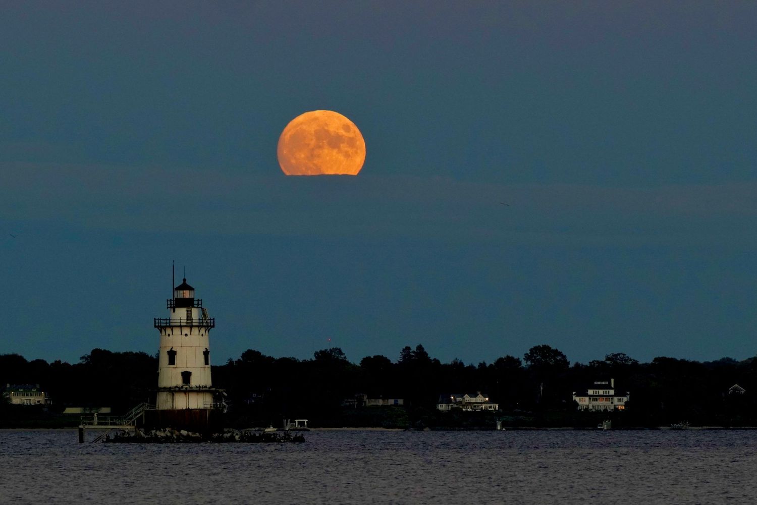 Bright full moon illuminates a lighthouse on the horizon over dark coastal waters.
