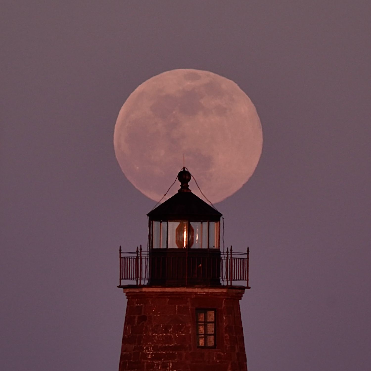 Full moon rises behind lighthouse silhouette creating dramatic dusk sky composition in pink and purple hues.