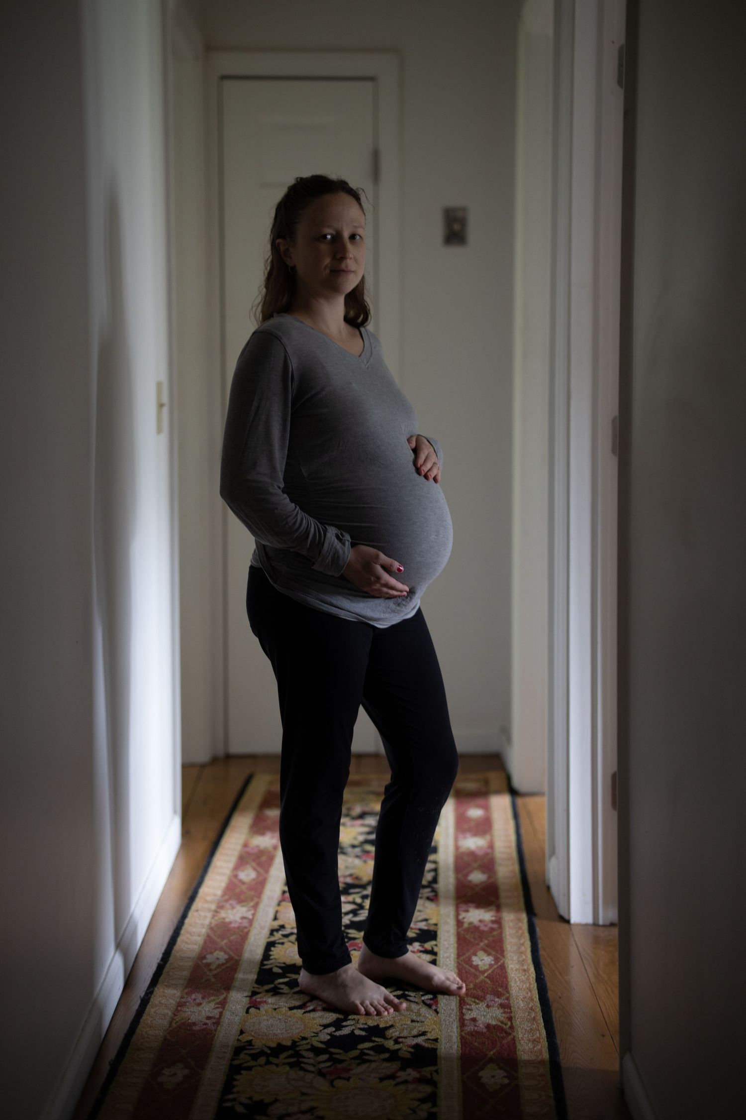 A pregnant woman stands in a dimly lit hallway wearing a gray sweater and black pants, standing on a patterned rug.