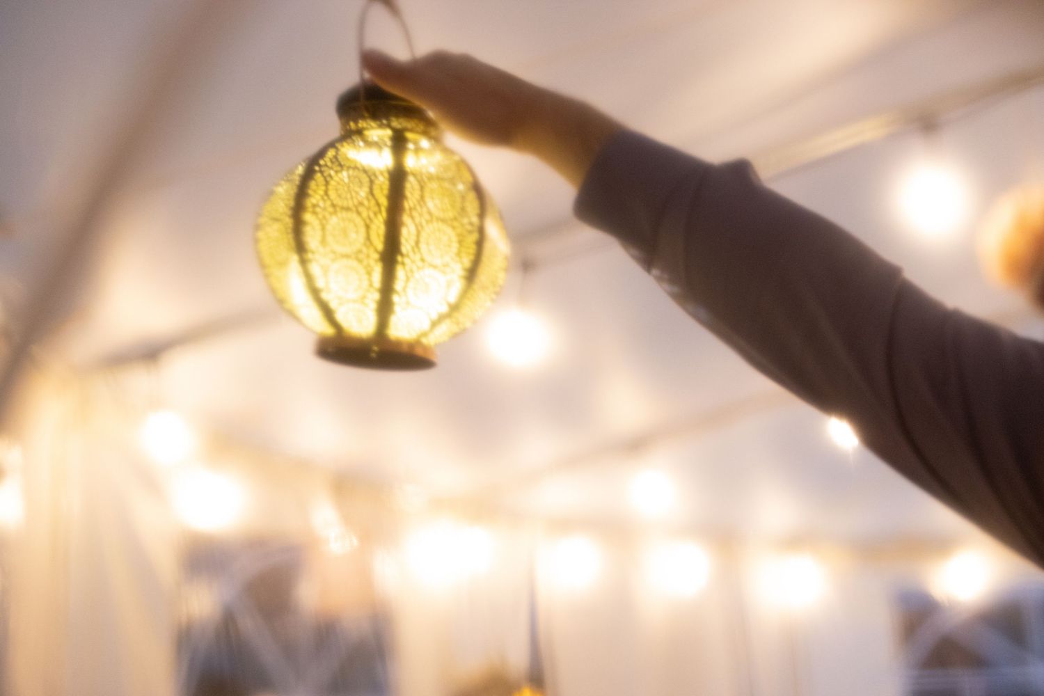 A hand reaches up to adjust a glowing paper lantern hanging from a string of decorative lights.
