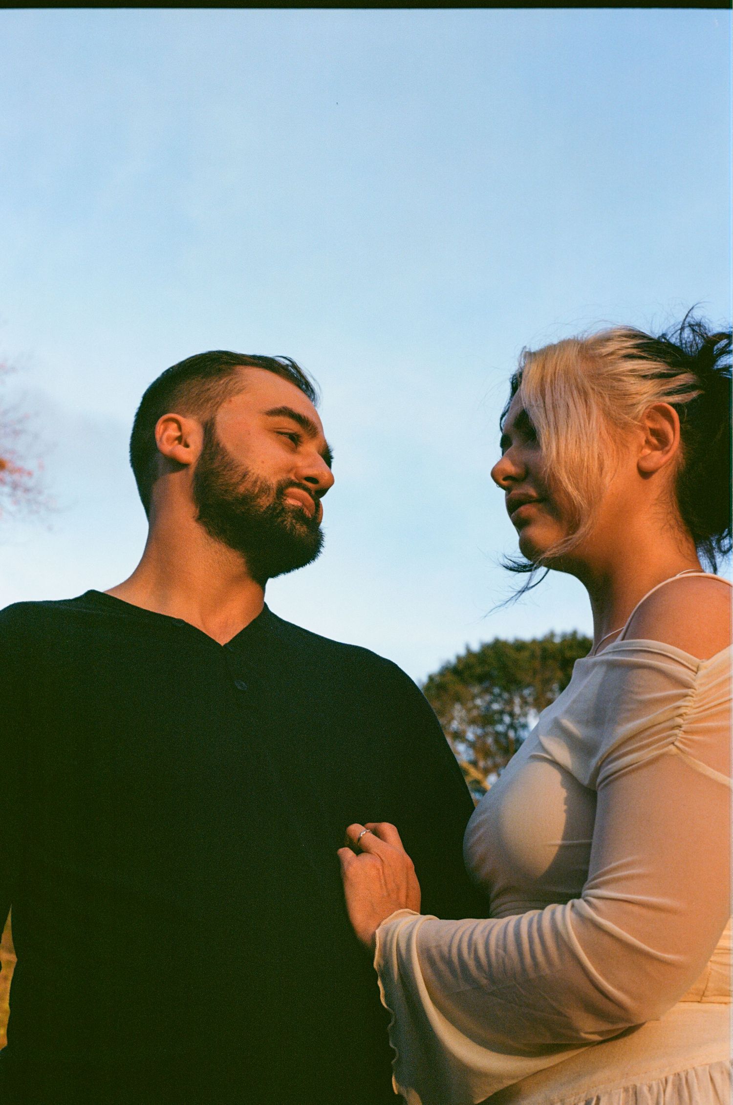 A bearded man in black and a blonde woman in white share an intimate moment outdoors under a blue sky.
